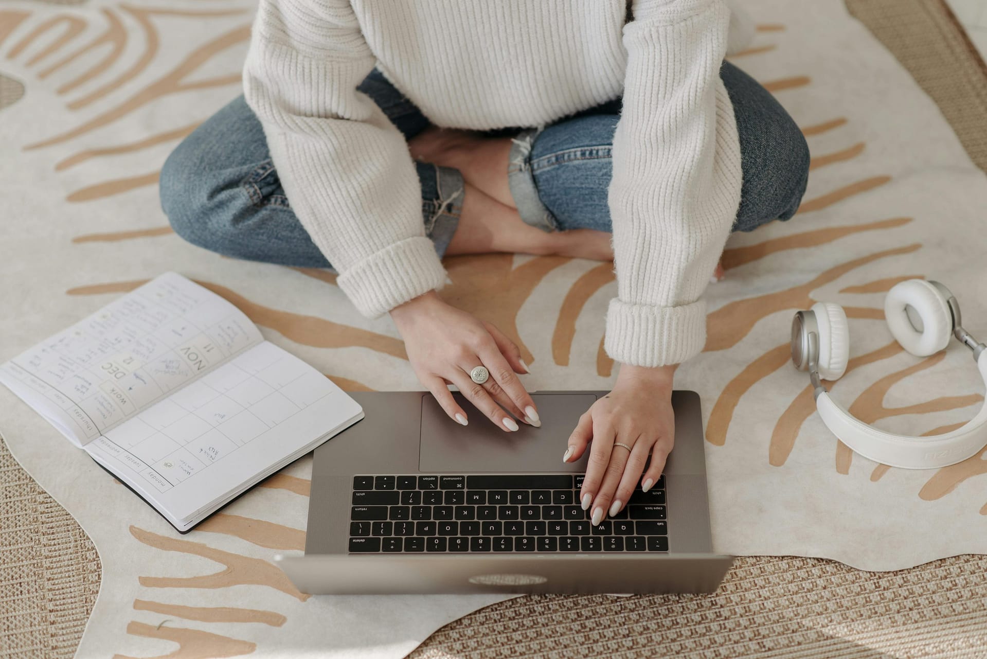 Girl sitting cross-legged on a rug working on a laptop, with headphones and an open diary beside her