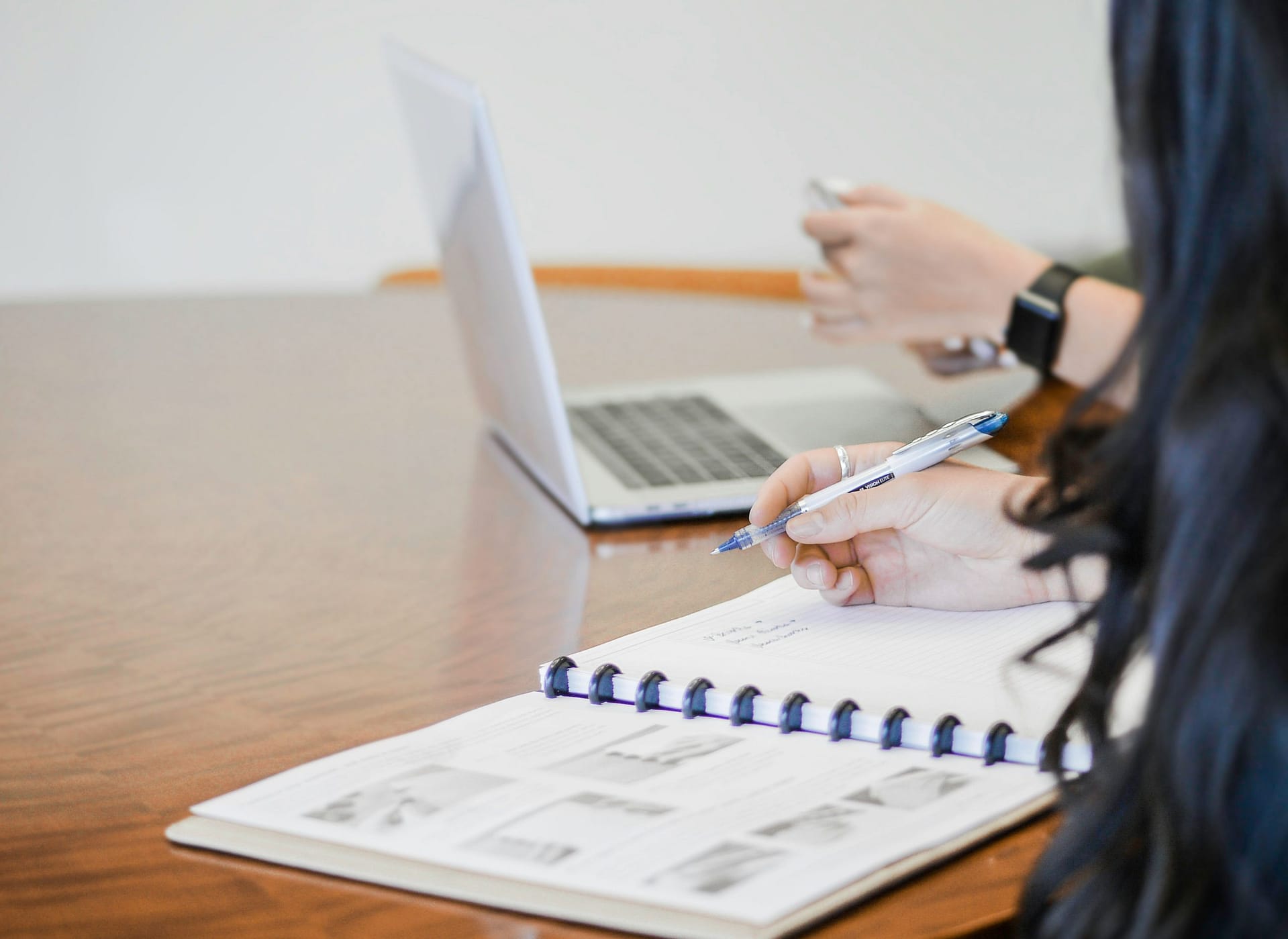 Two students working at a desk, one with an open book, one on a laptop