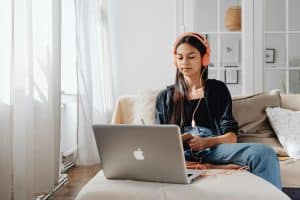 University-age student studying on a couch with a laptop and headphones by a window